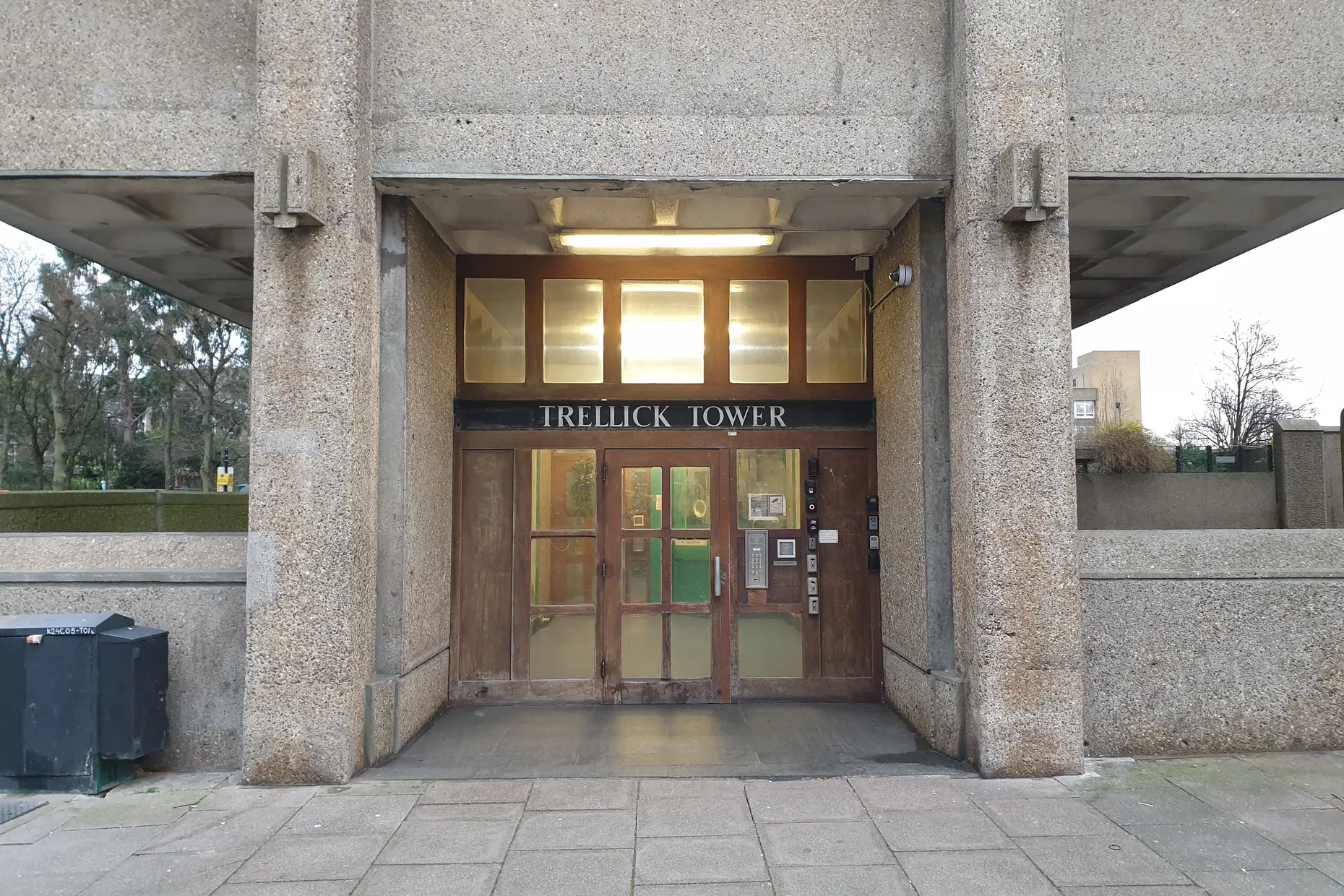 The entrance to Trellick Tower, board-marked concrete columns framing a wooden door under a lit canopy