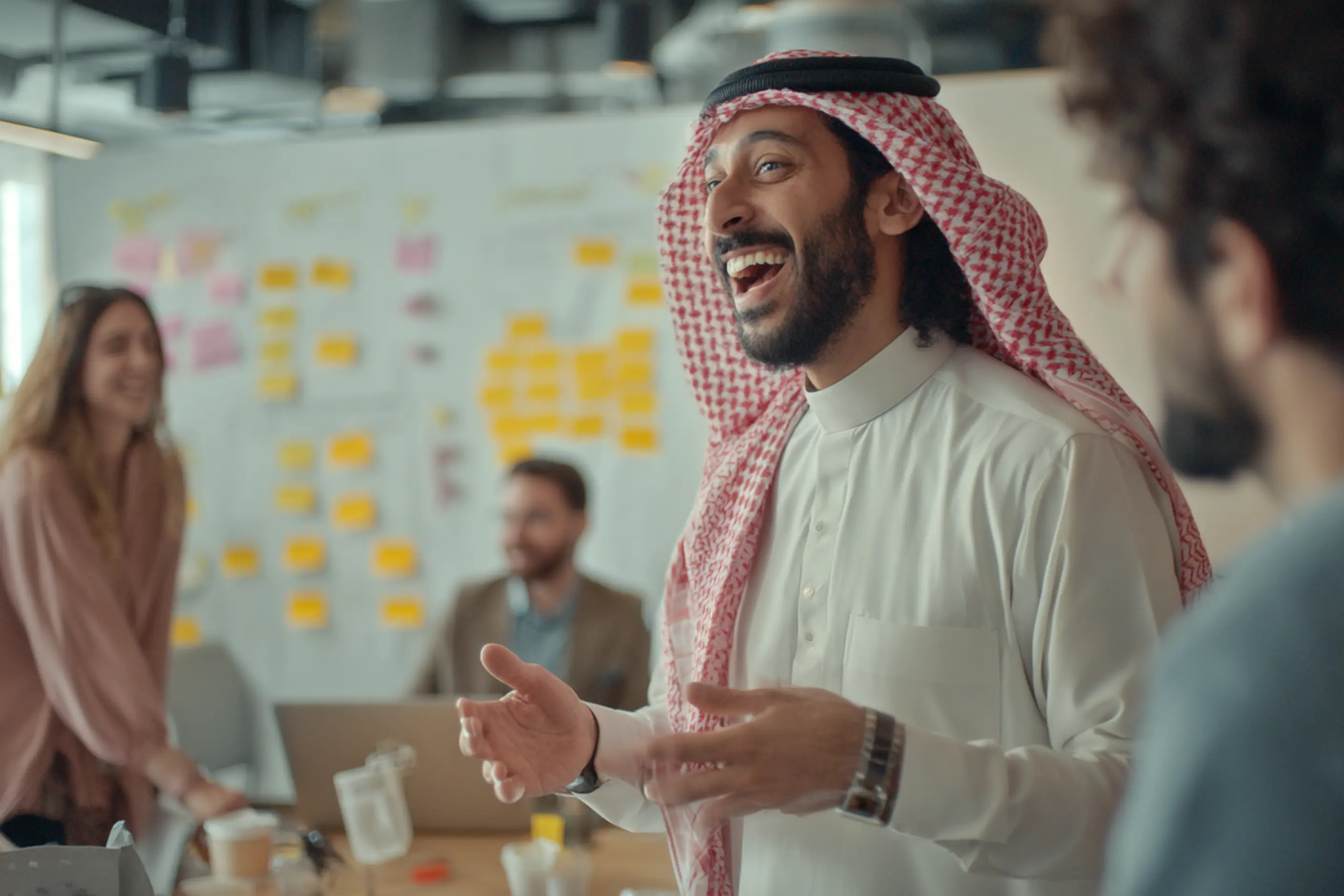 A laughing man in a red keffiyeh gestures as he talks in a workshop room, colleagues and a sticky-note wall behind him