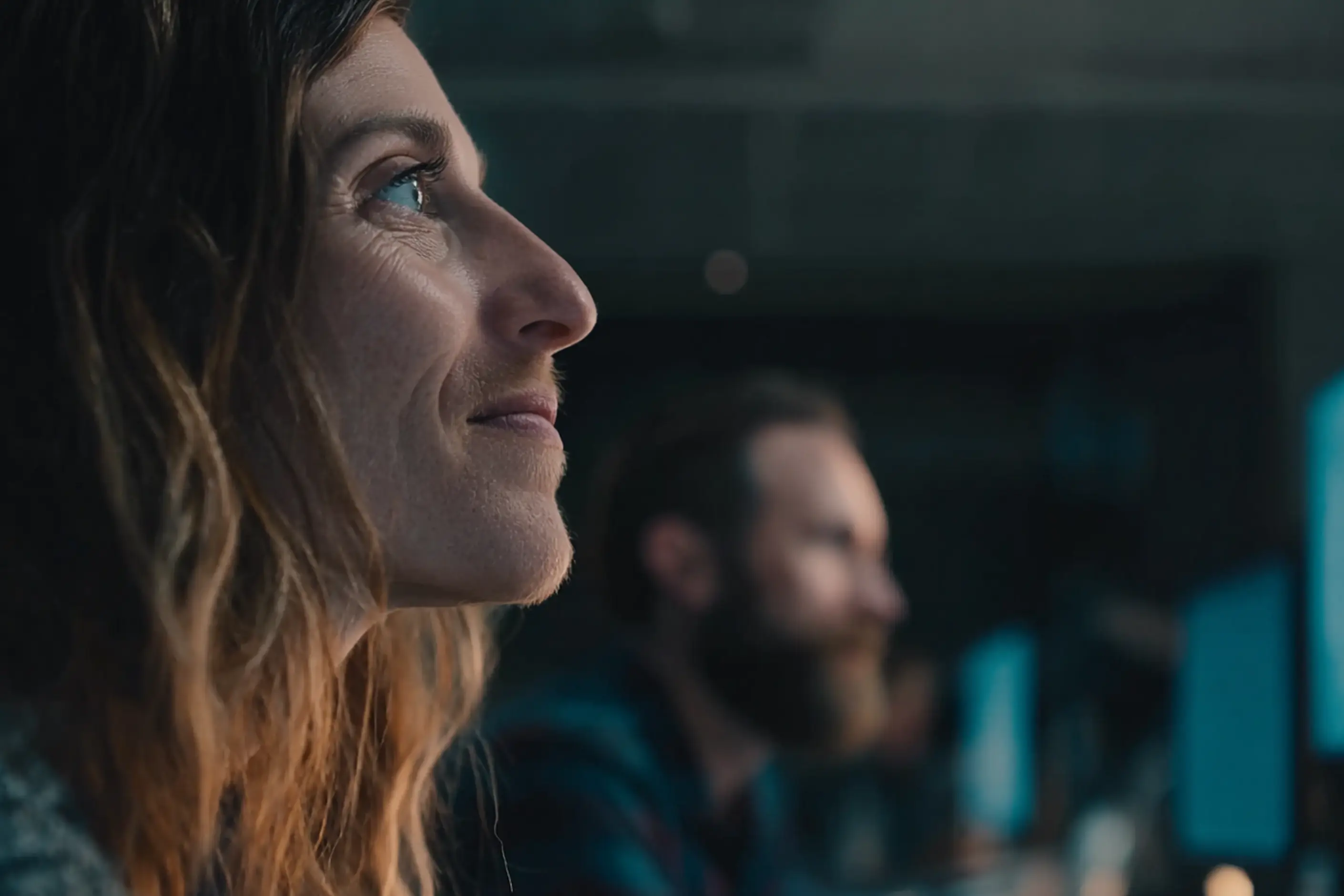 A woman in profile watching a session intently, a colleague blurred beside her at a screen