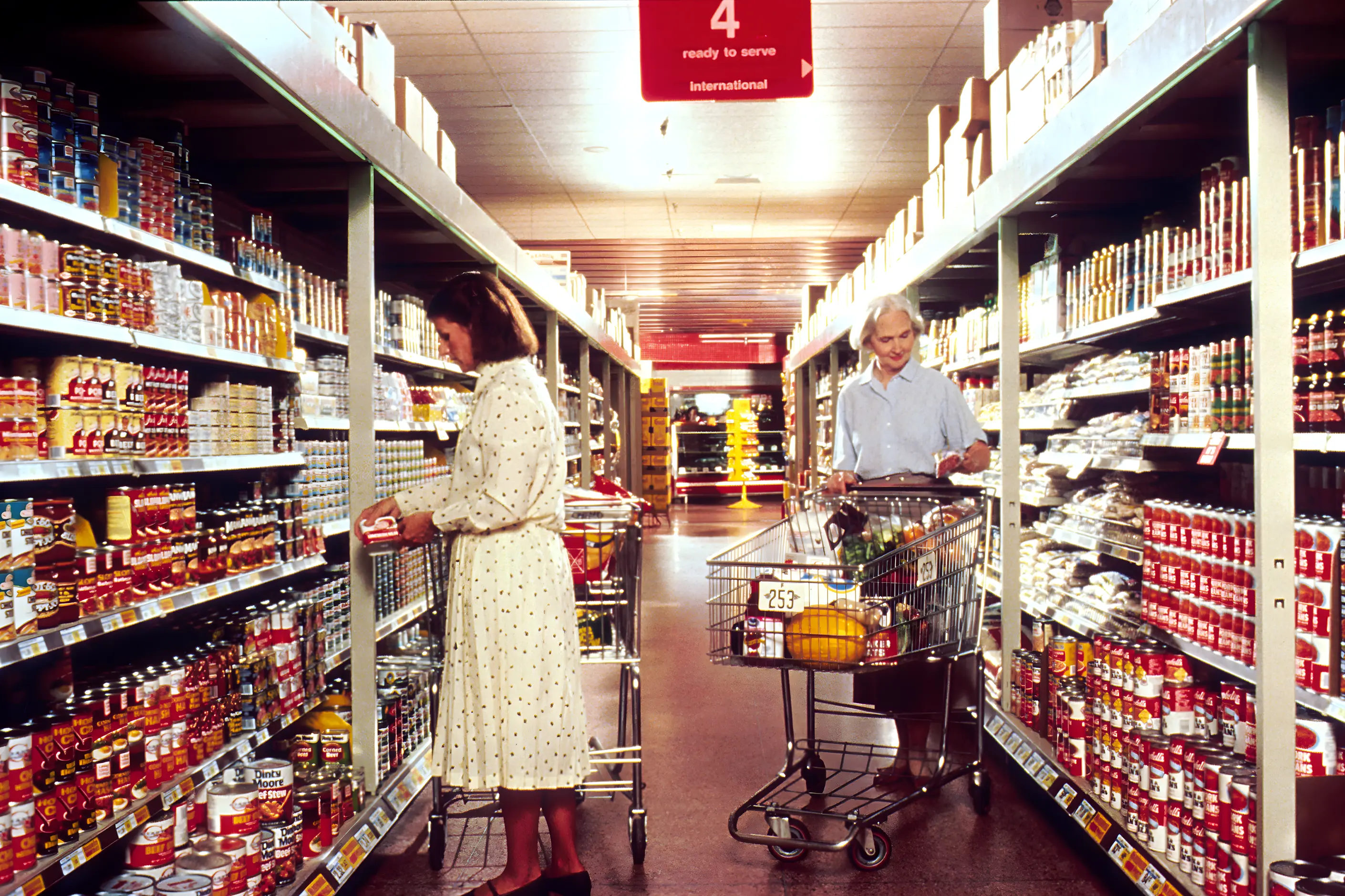 Two shoppers in a supermarket aisle lined floor to ceiling with branded cans and packets