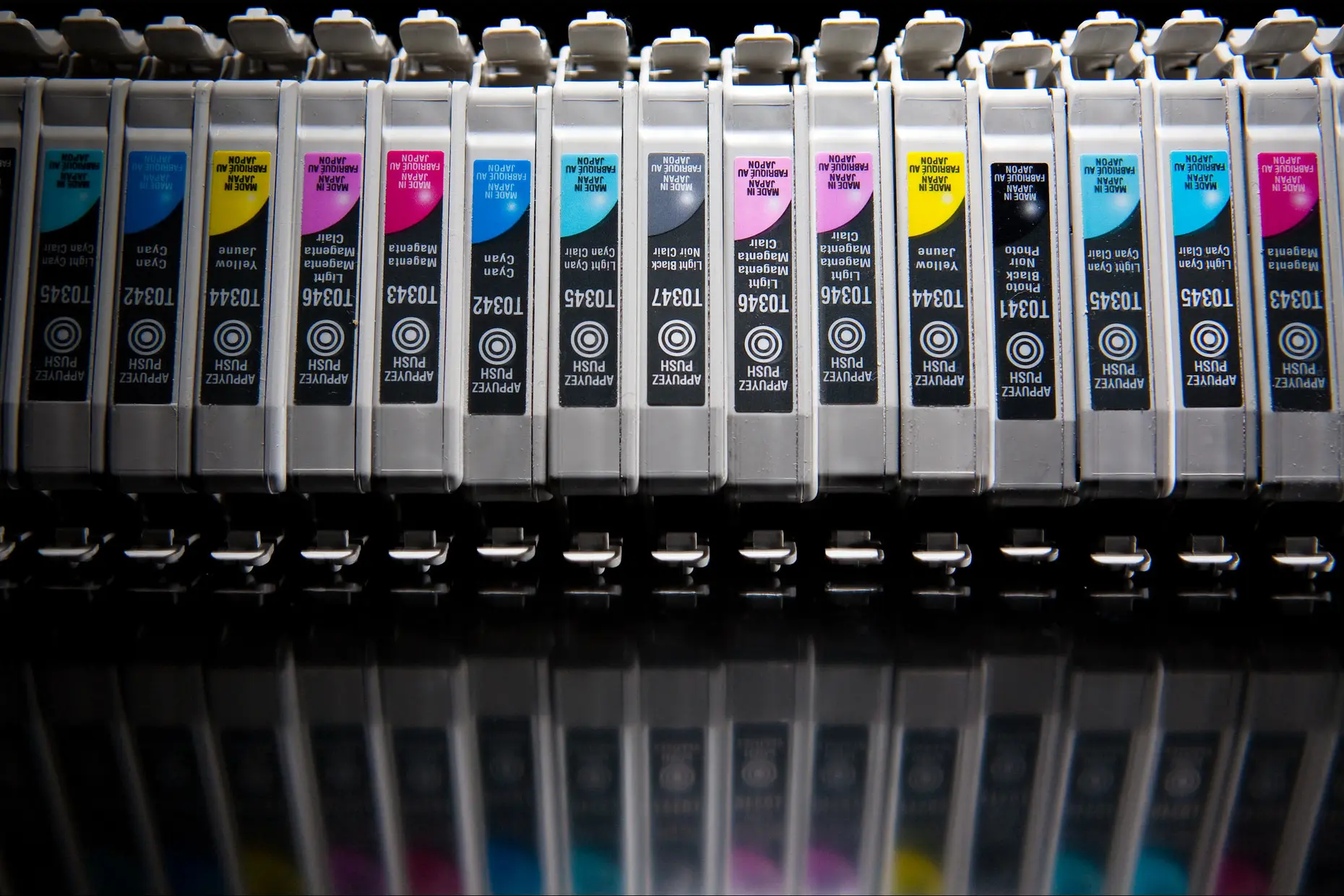 A row of cyan, magenta, yellow and black printer ink cartridges lined up, reflected below