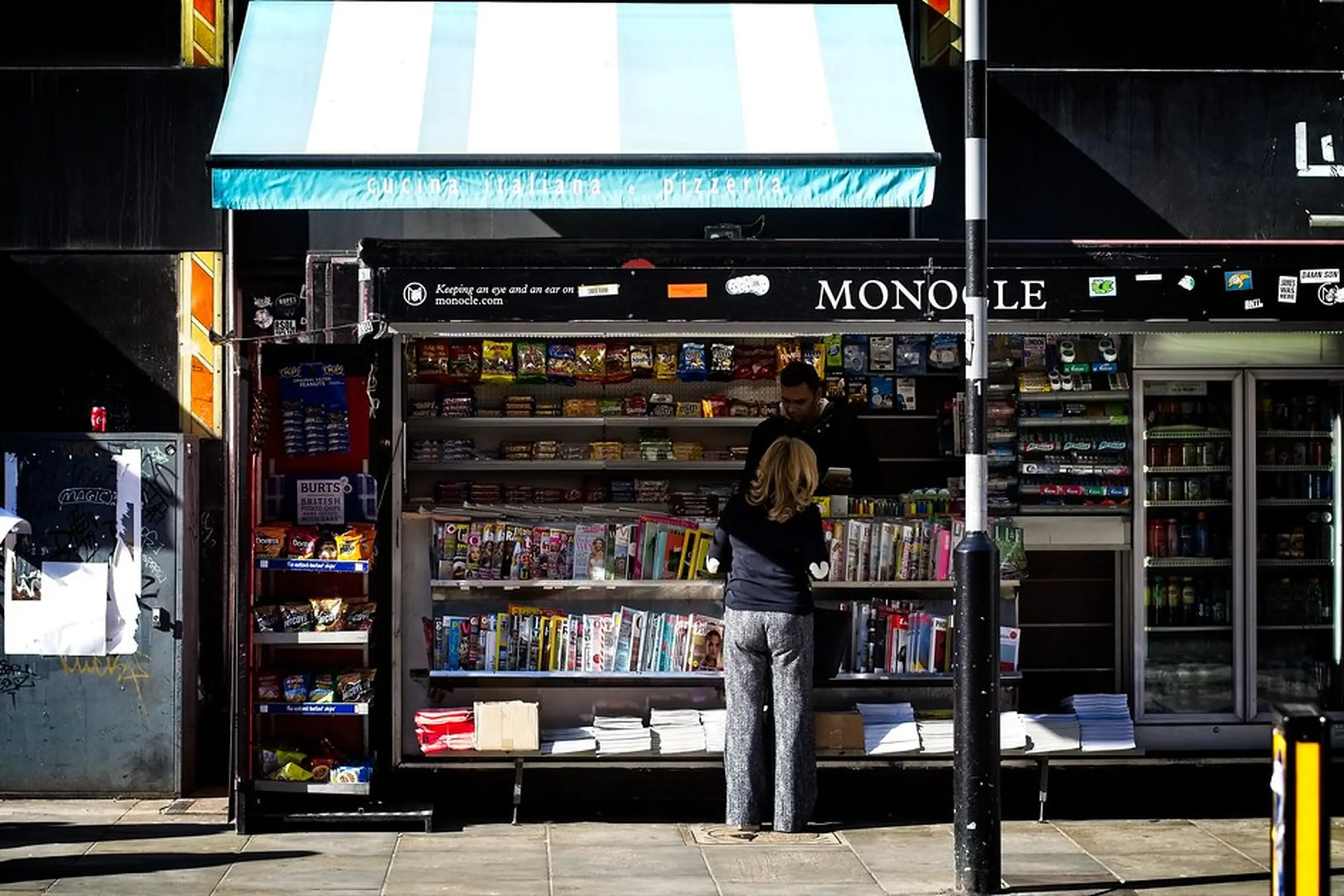 The Monocle kiosk on a London street, its racks of magazines and books under a striped awning, a reader browsing
