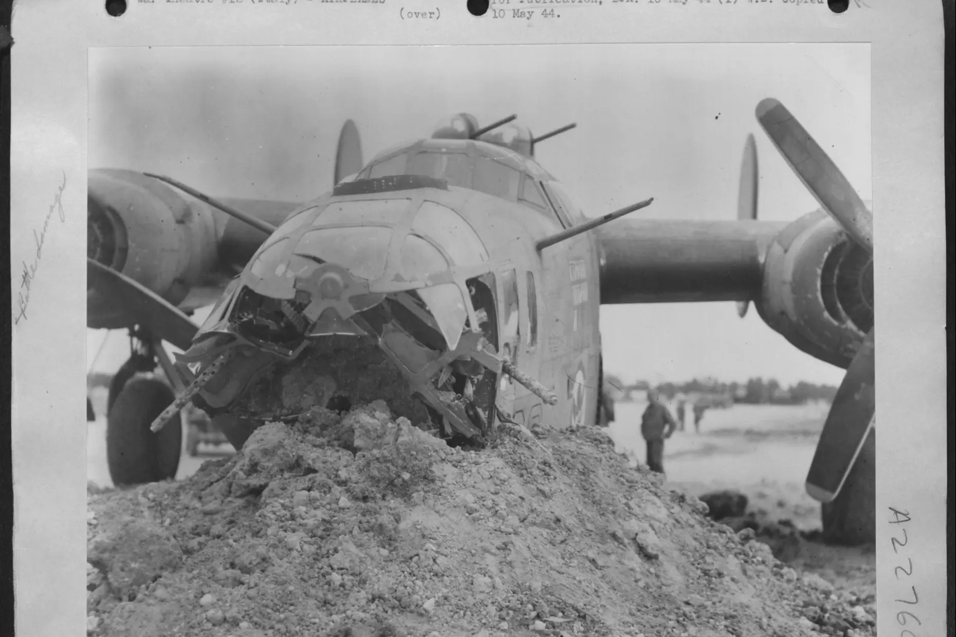 A wartime press print of a B-24 bomber with its nose shattered by flak, resting on a mound of earth after a crash landing