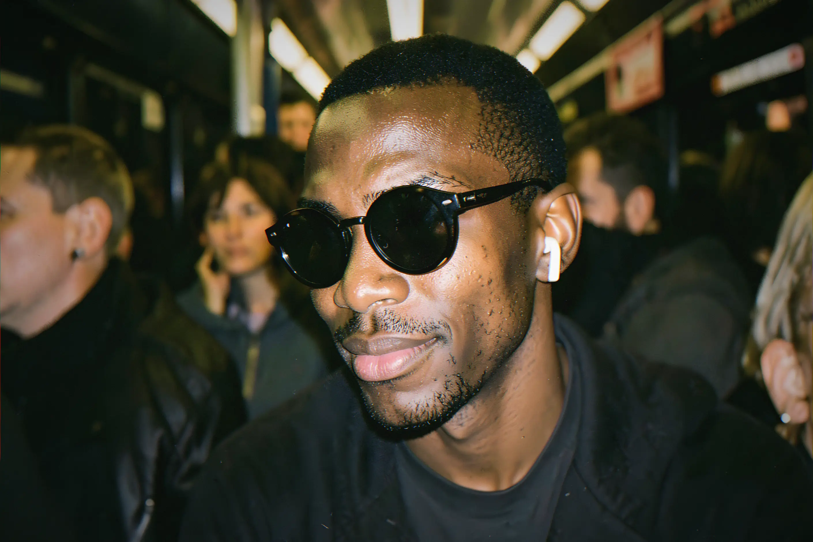 A young blind man in dark round glasses and a single earbud smiles as he listens, in a crowded late-night train carriage