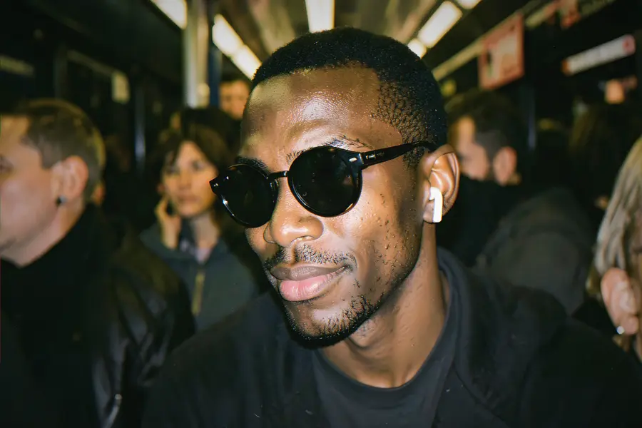 A young blind man in dark round glasses and a single earbud smiles as he listens, in a crowded late-night train carriage
