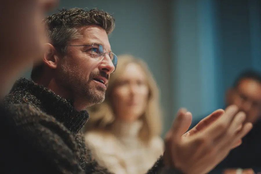 A bearded man in glasses gestures as he speaks during a small focus group session around a table
