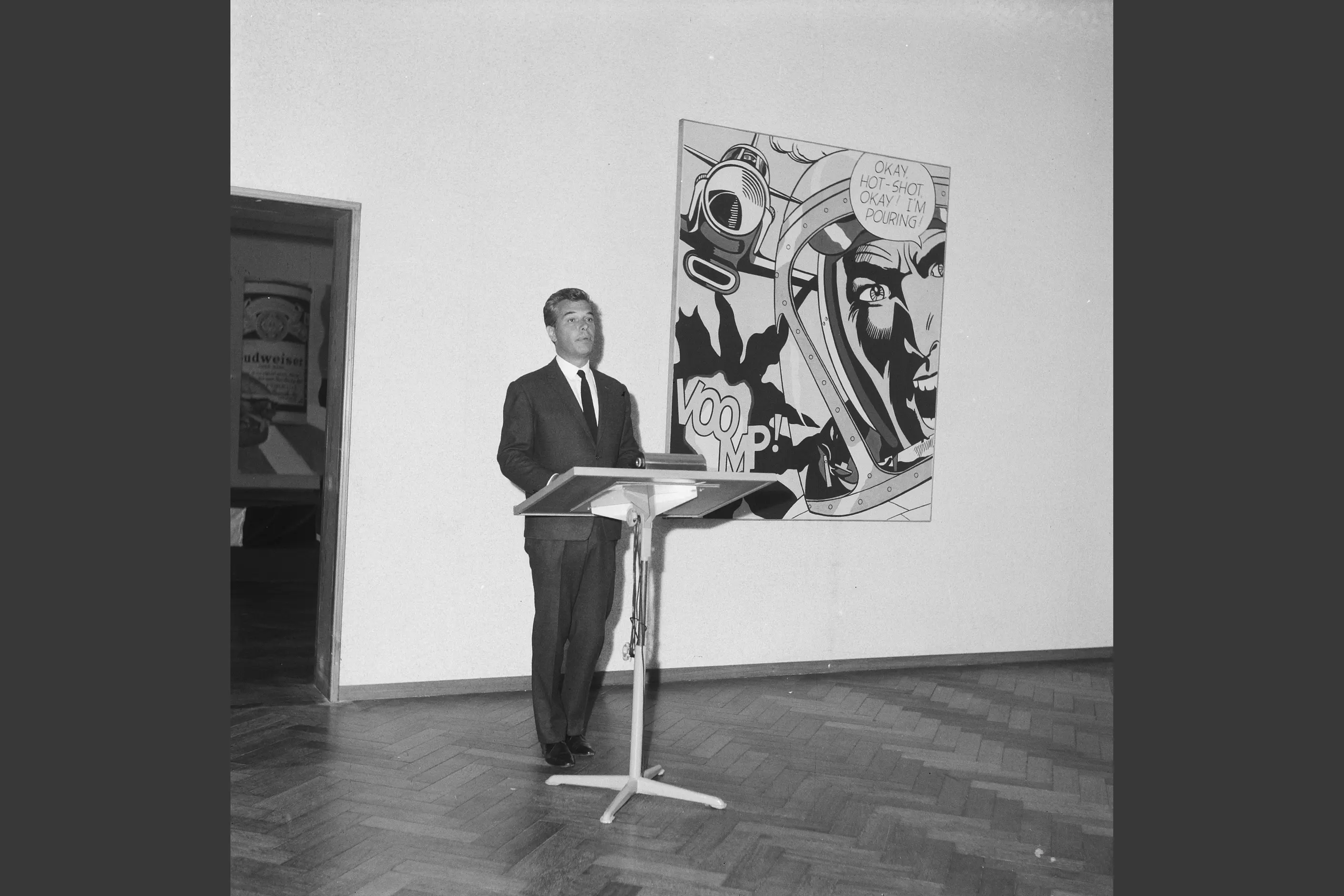 A man speaks at a lectern beside a large Lichtenstein comic panel with a speech bubble and VOOM lettering