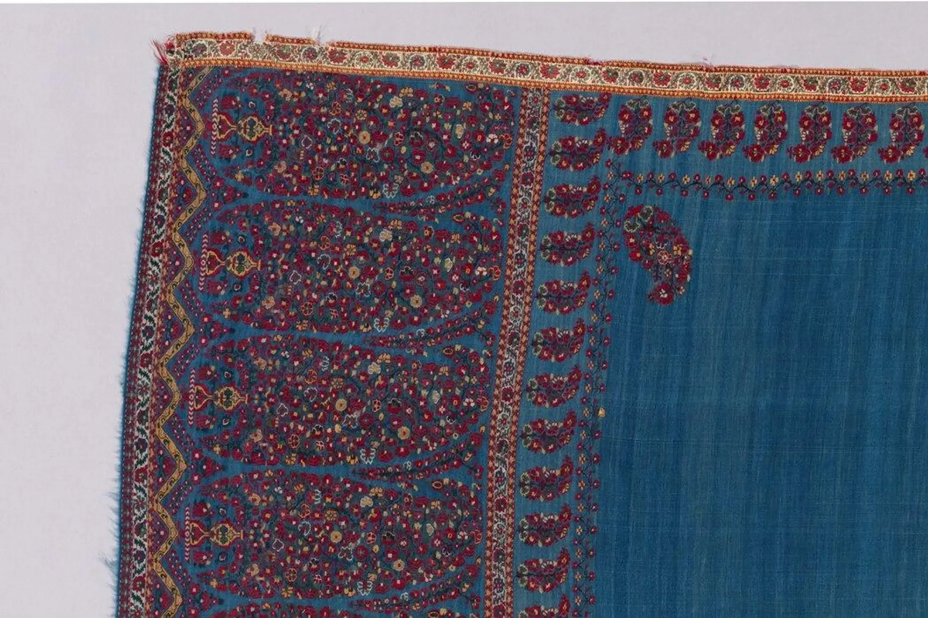 Close up corner of a woven shawl showing dense paisley boteh motifs in red and gold on blue