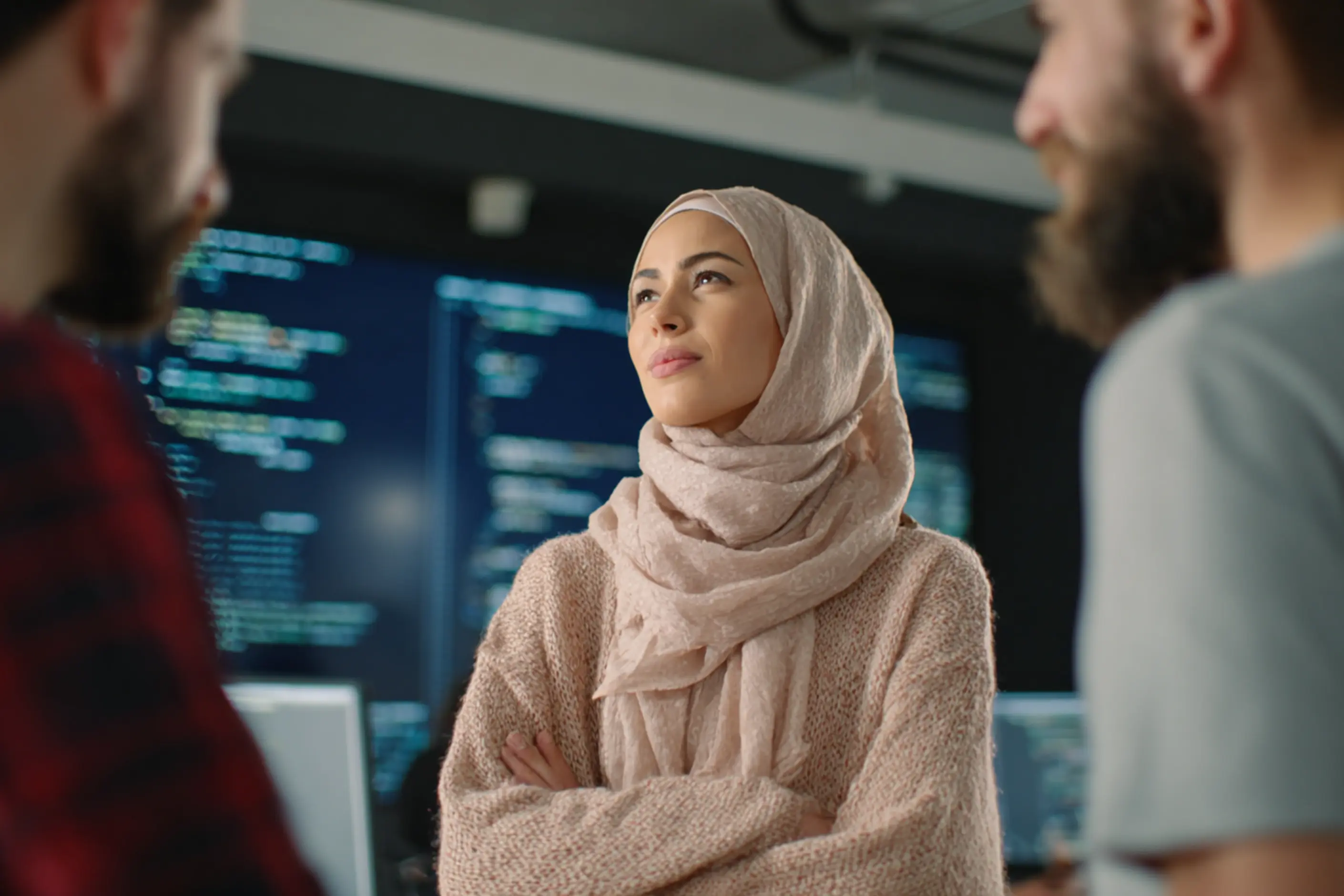 A woman in a pink hijab stands with folded arms among teammates in front of code screens