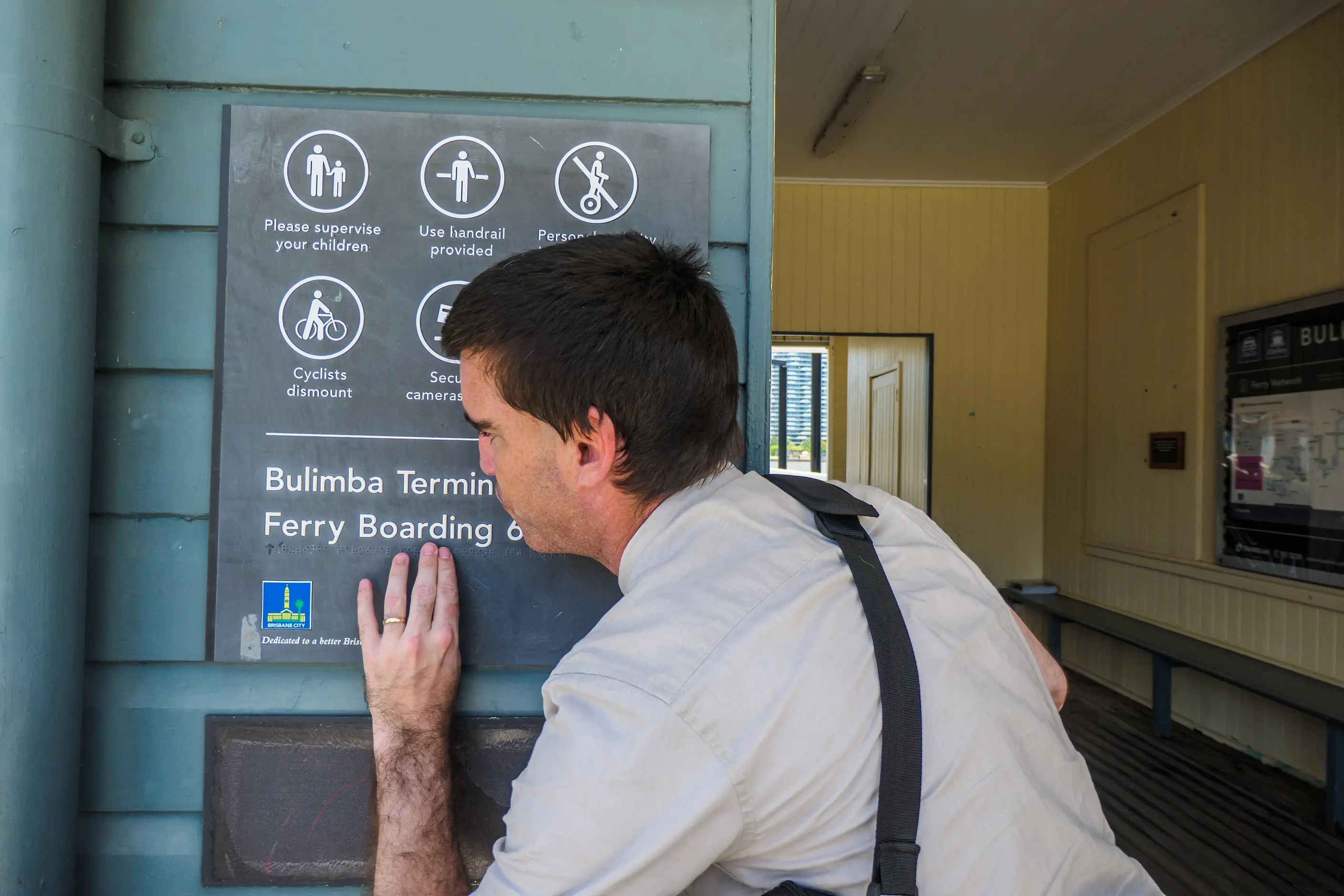 A man reading a tactile wayfinding sign by touch at a ferry terminal, raised pictograms and braille on a dark panel