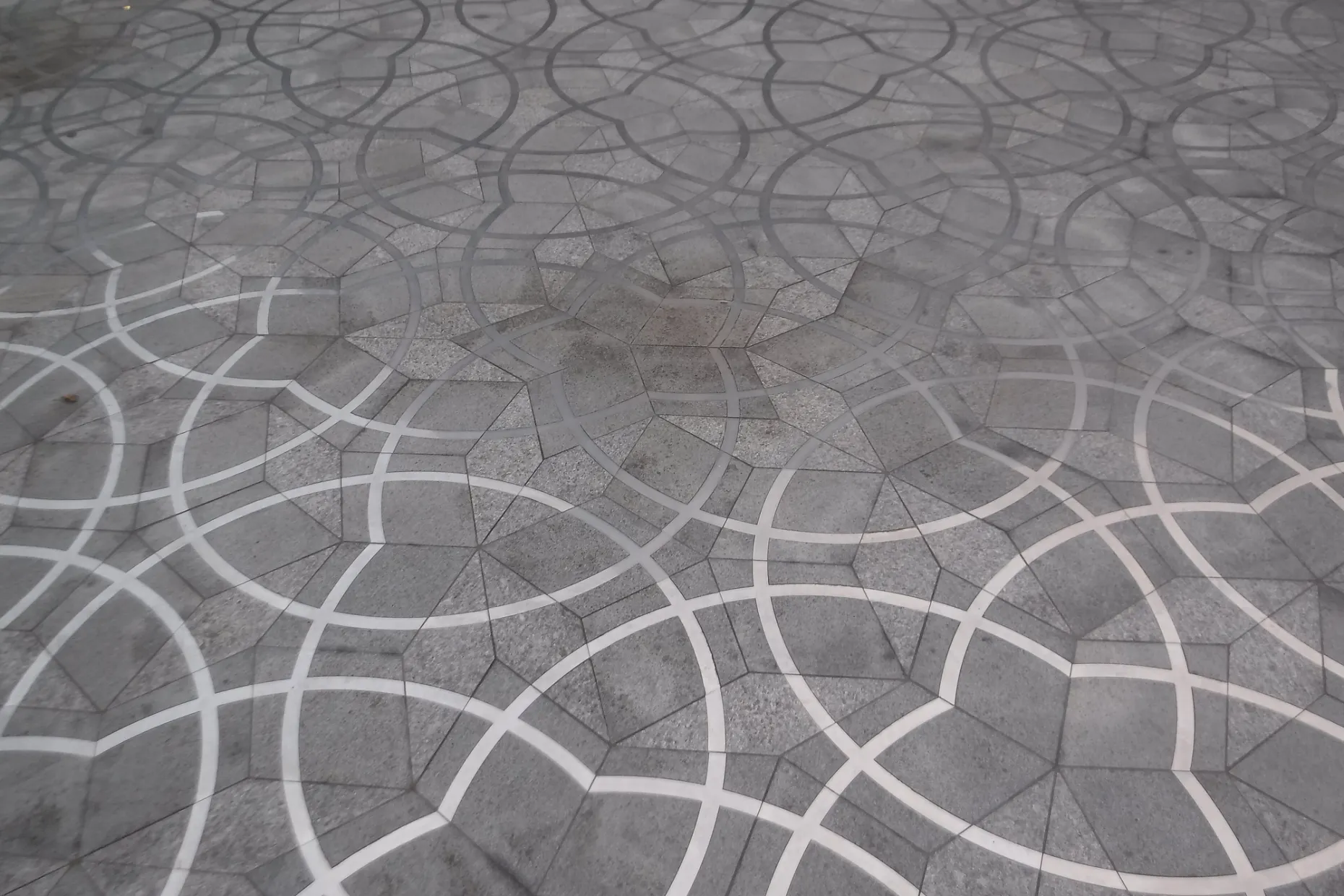 A grey stone Penrose tiling paving a plaza, white inlaid arcs running across kites and darts that never repeat