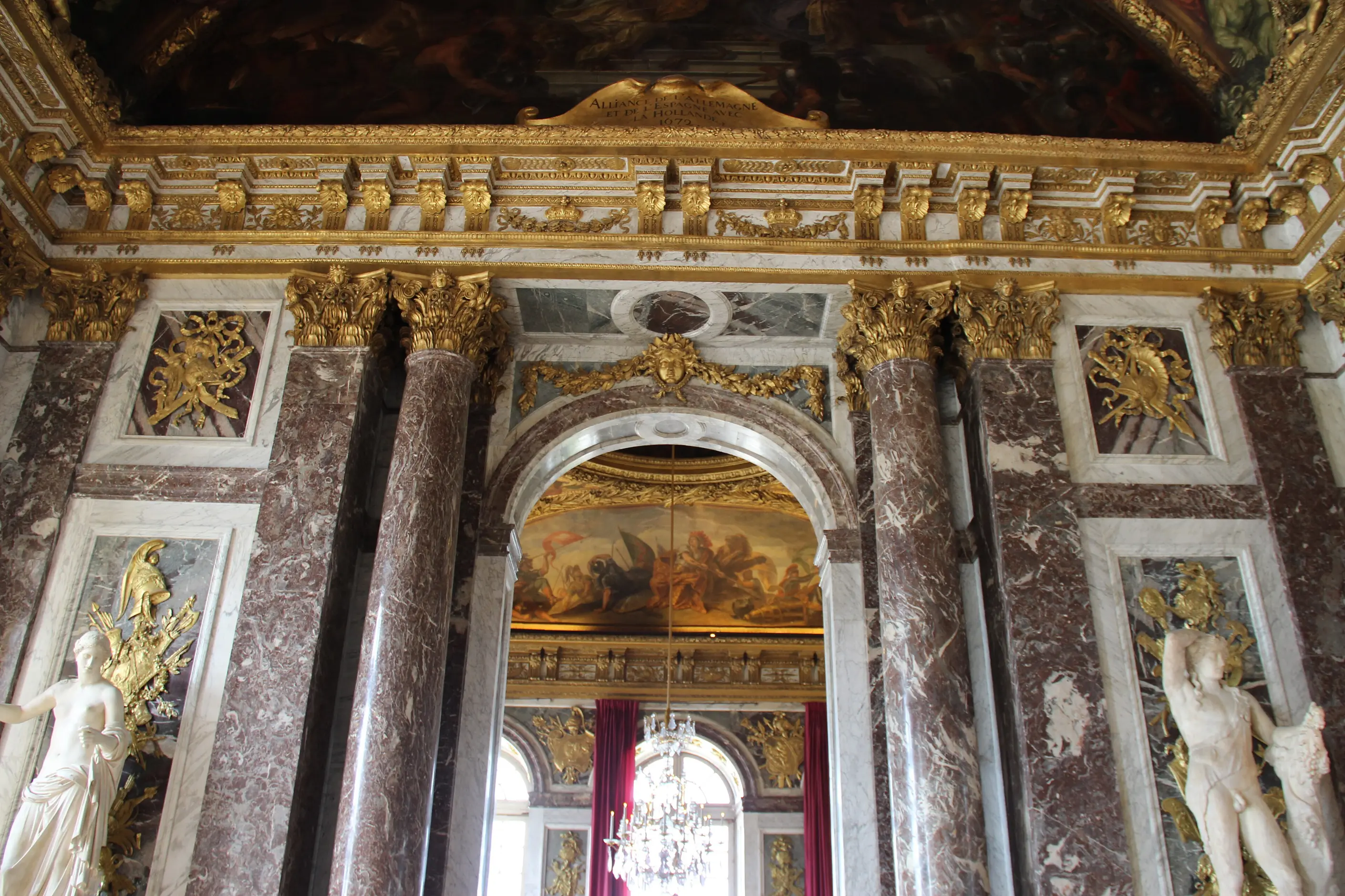Red marble columns with gilded capitals, trophies and white statues framing an arched doorway at Versailles