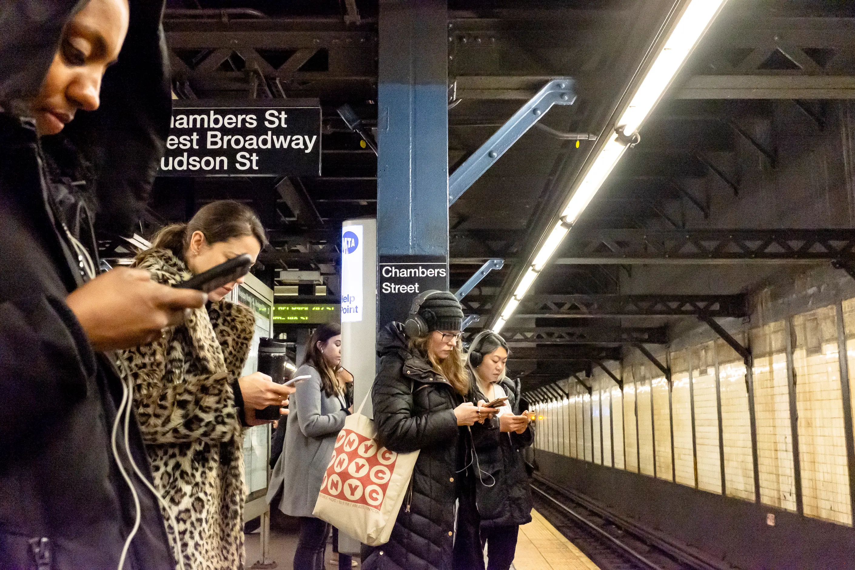 Commuters on a New York subway platform, each looking down at a phone while waiting for a train