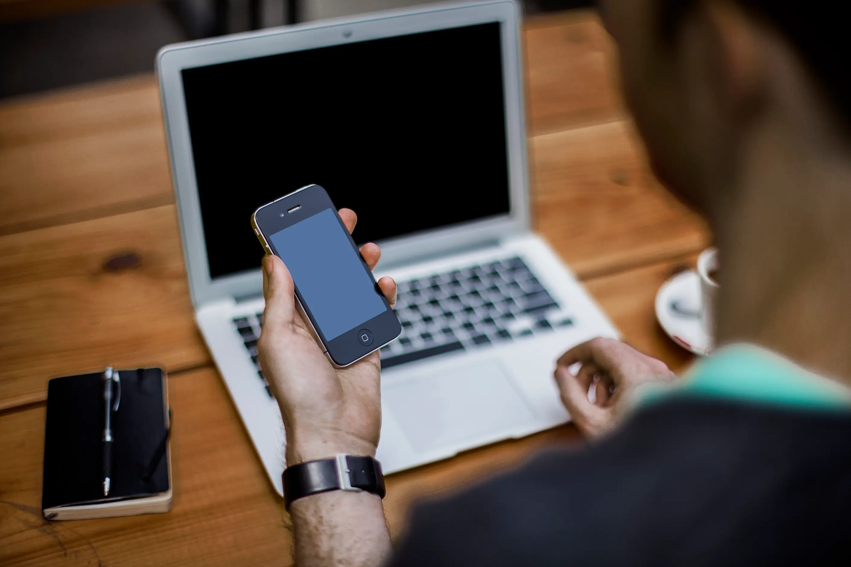 A person at a wooden desk holding a smartphone over an open laptop, a notebook beside them