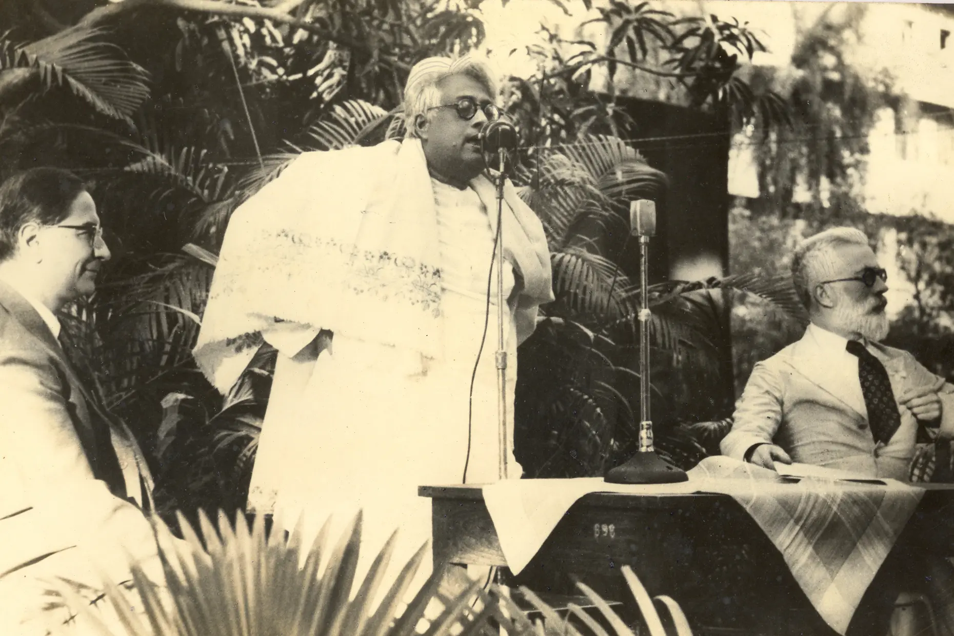 Three men at an outdoor lectern, one in white robes speaking into a microphone, a bearded man seated at right