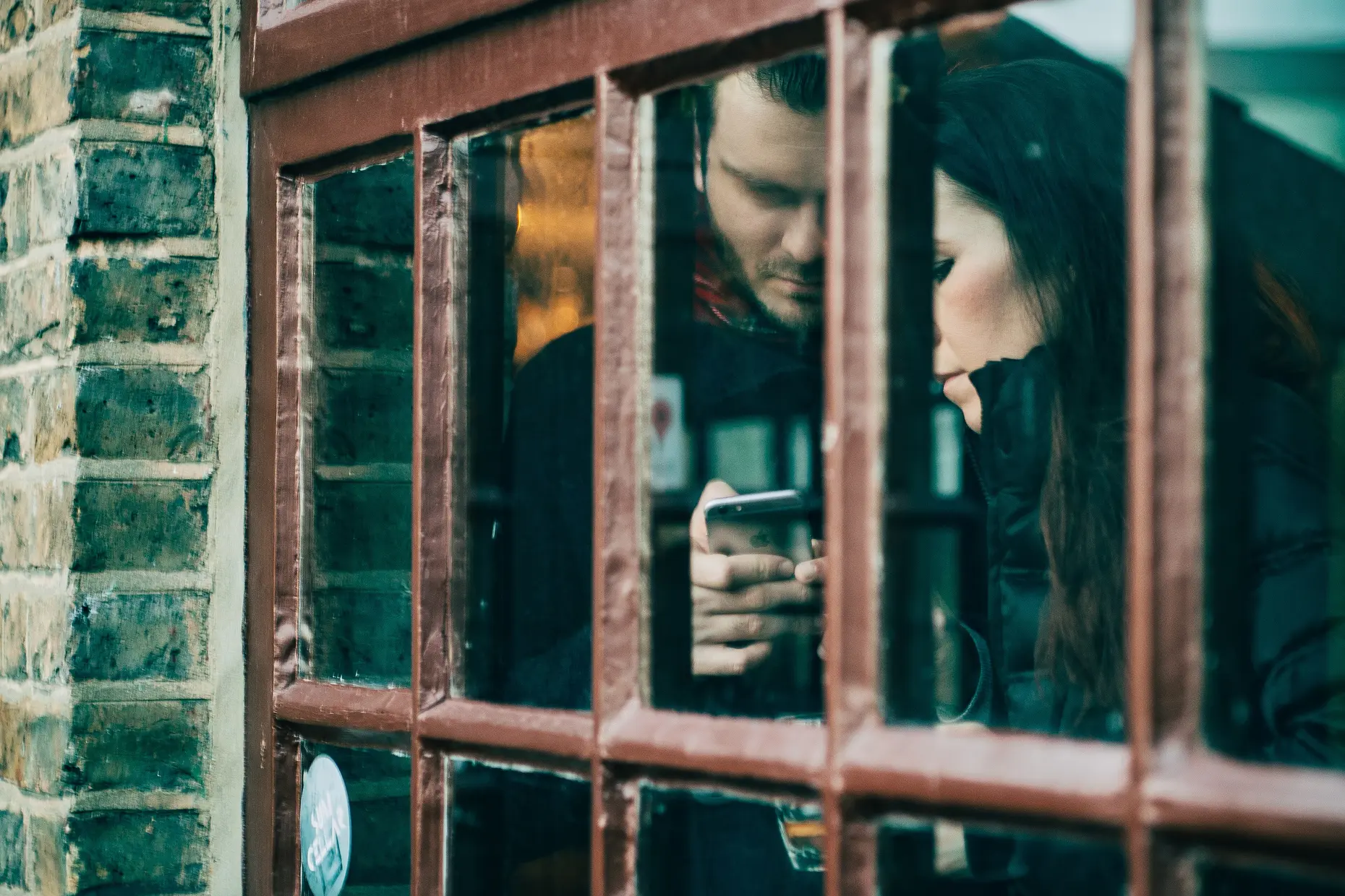 Two people seen through an old shop window, each looking down at their own phone