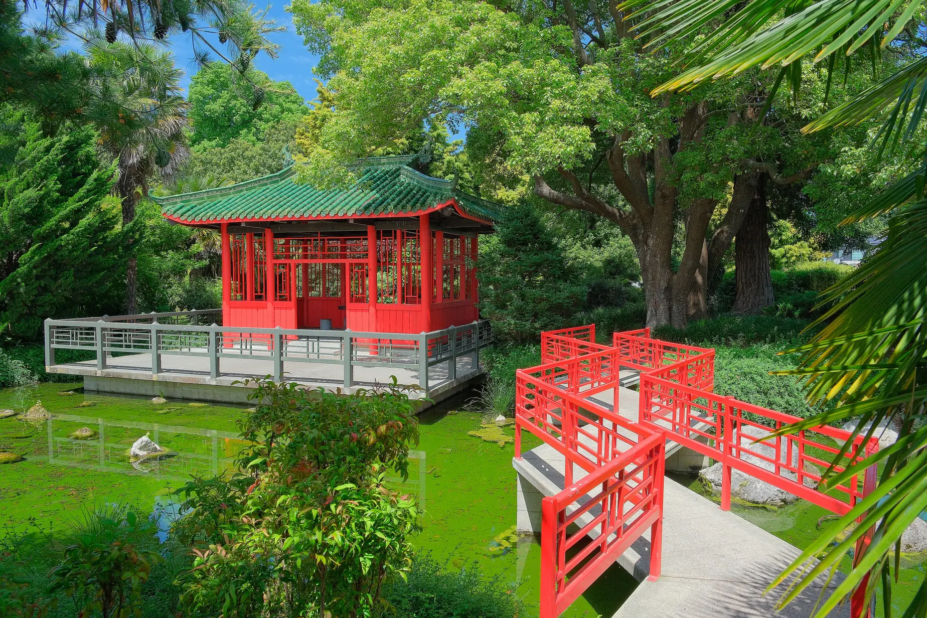 A red zigzag bridge crossing a green pond towards a red pavilion with a green tiled roof