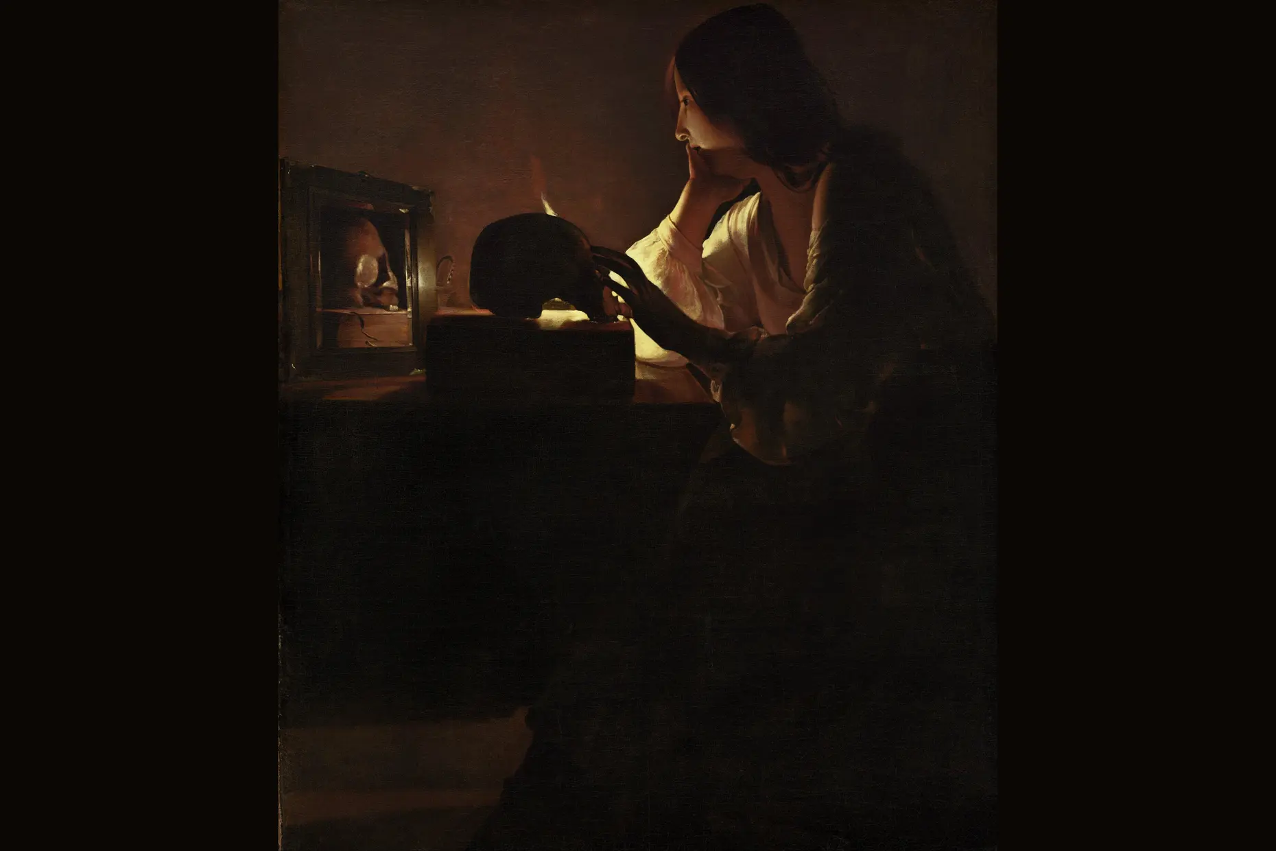 Woman lit by a single candle flame resting her hand on a skull in a dark room